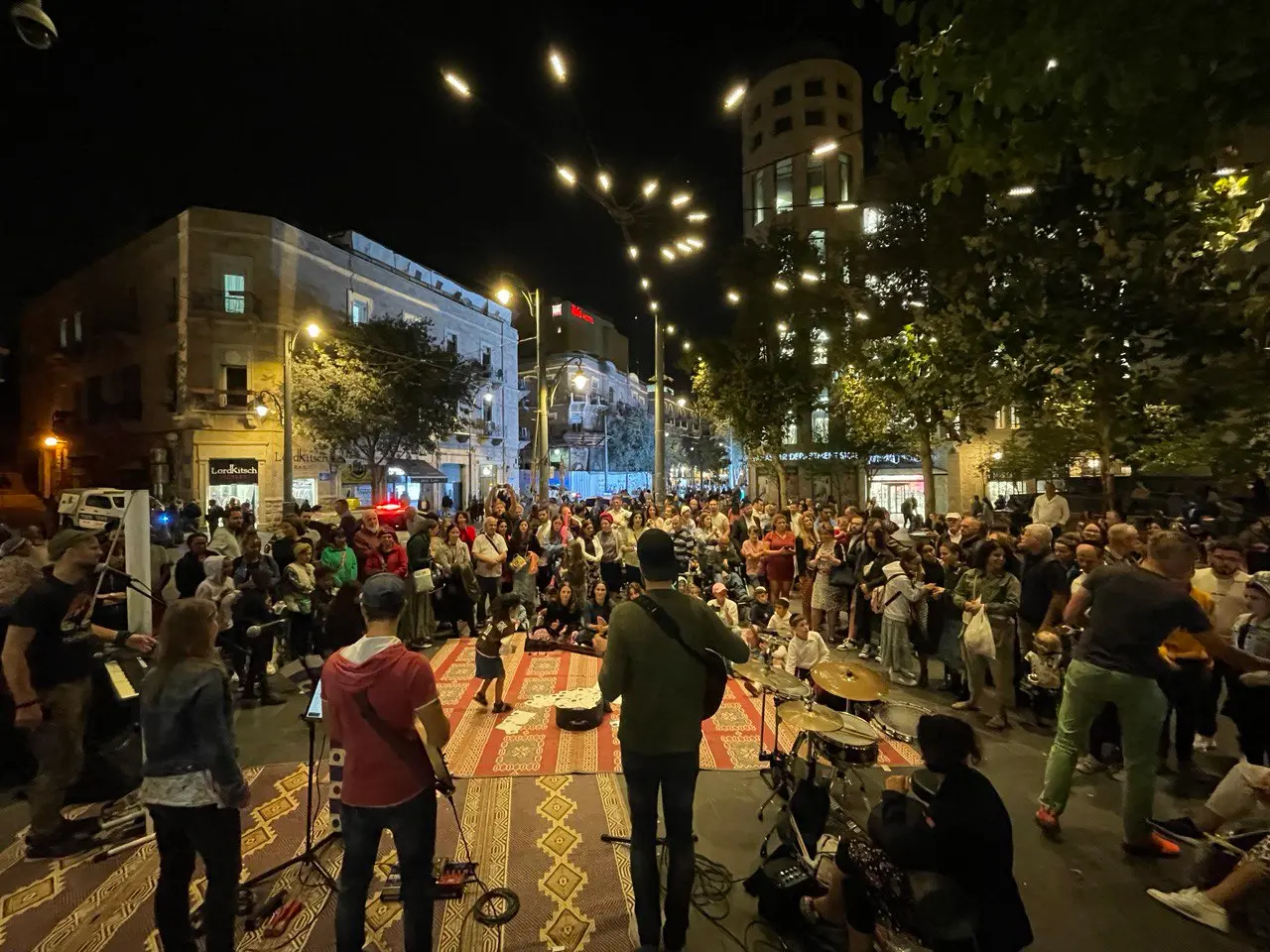 Blick von hinter der Band über den Teppich auf die Zuhörer, dahinter die nächtliche Straße mit Lichterketten