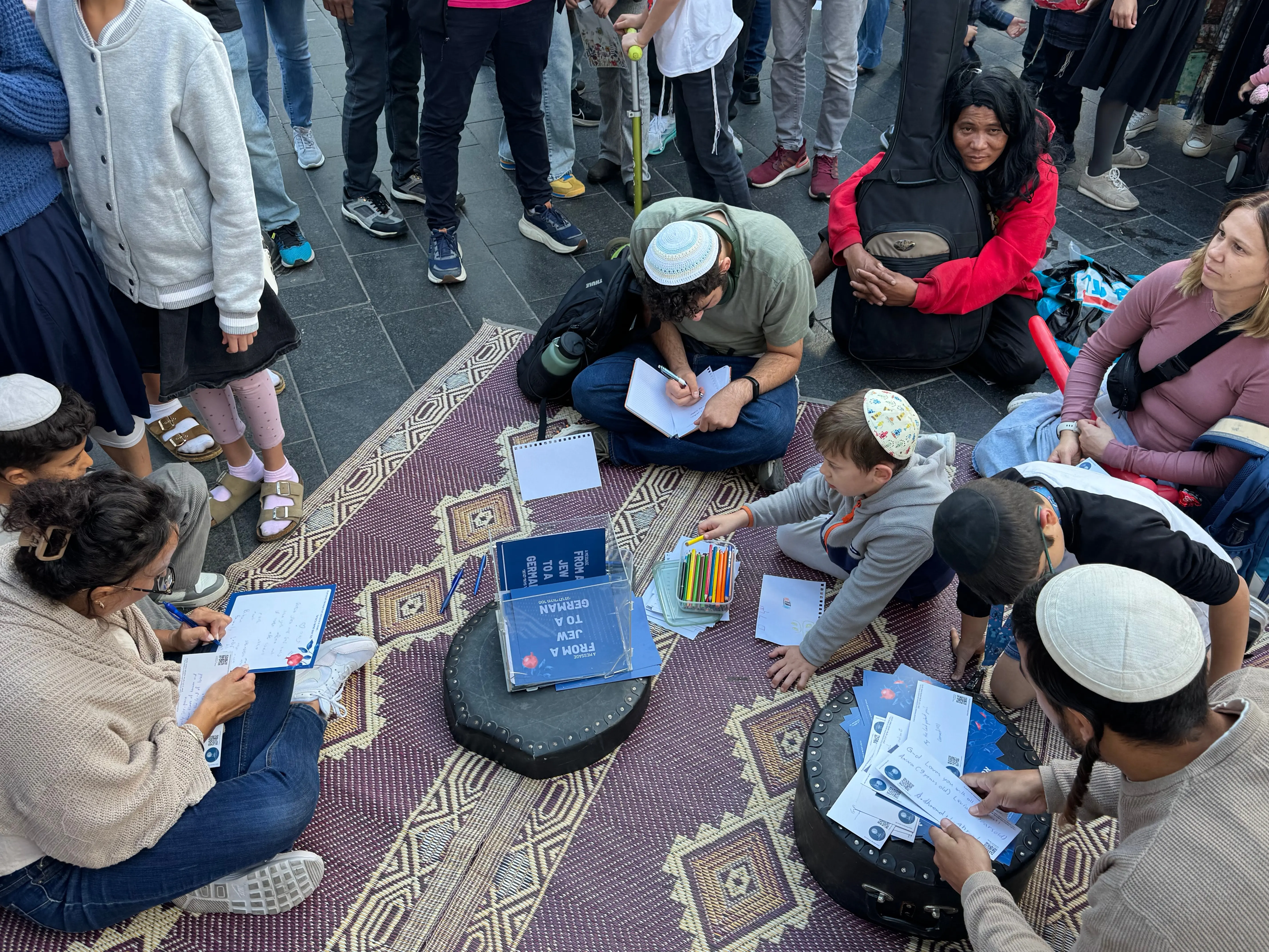 Blick von oben auf den Teppich in der Fußgängerzone: Menschen sitzen im Kreis und schreiben Postkarten, in der Mitte der Aufsteller „A message from a Jew to a German“